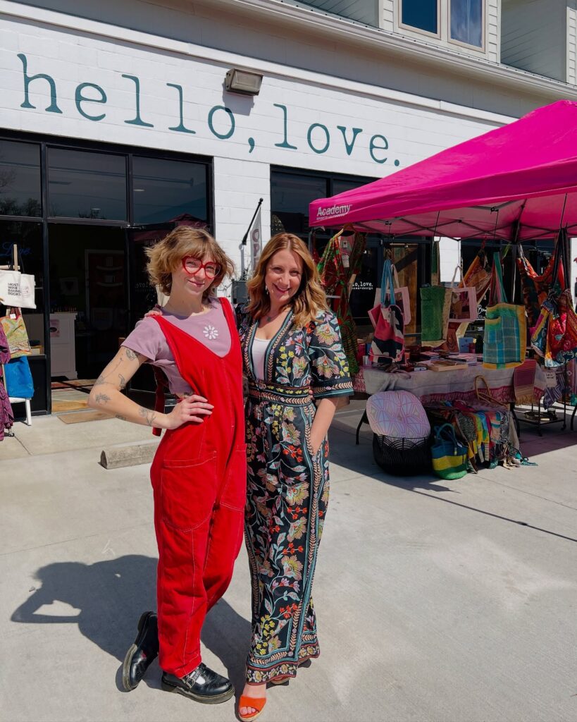 Two women wearing colorful and fashionable clothing standing out front of hello, love boutique. Table with boutique clothing and goods on a table.