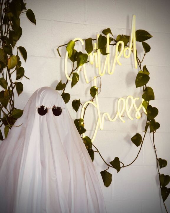 Person in a sheet and sunglasses posing like a ghost outside in the Cargo District by an LED sign for grilled cheese and hanging vines. 